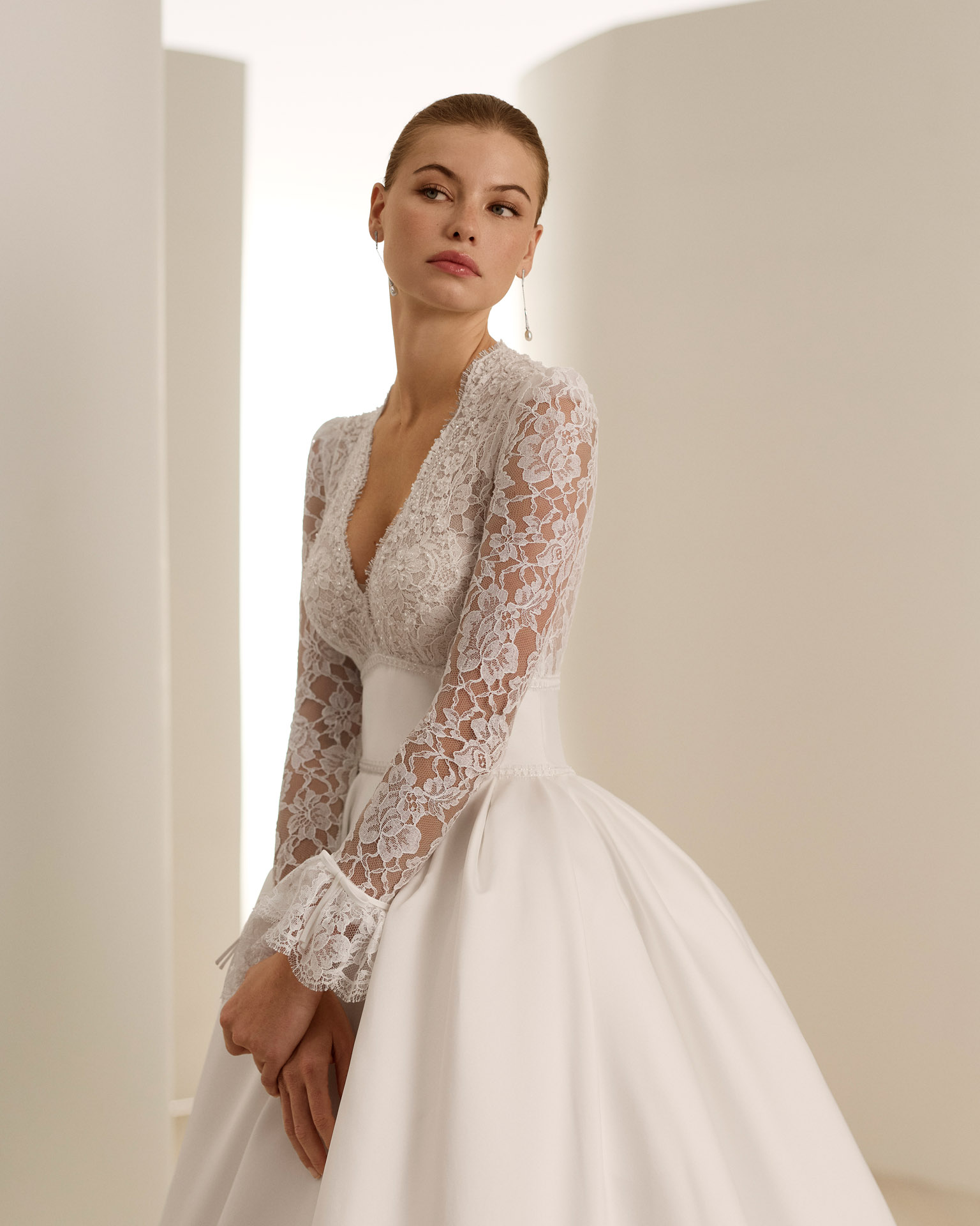 DARIA Abito da sposa silhouette principessa con scollatura profonda. Corpetto in pizzo ricamato con ornamenti di strass, gonna in mikado Magnolia. Maniche lunghe con volant e schiena abbottonata.